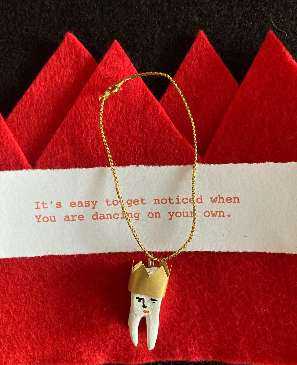 A christmas cracker made from red felt, with a piece of paper running across it that says 'It's easy to get noticed when you are dancing on your own'. A small model of a molar tooth wearing a gold crown hanging from it sits, on top of the red felt crown.