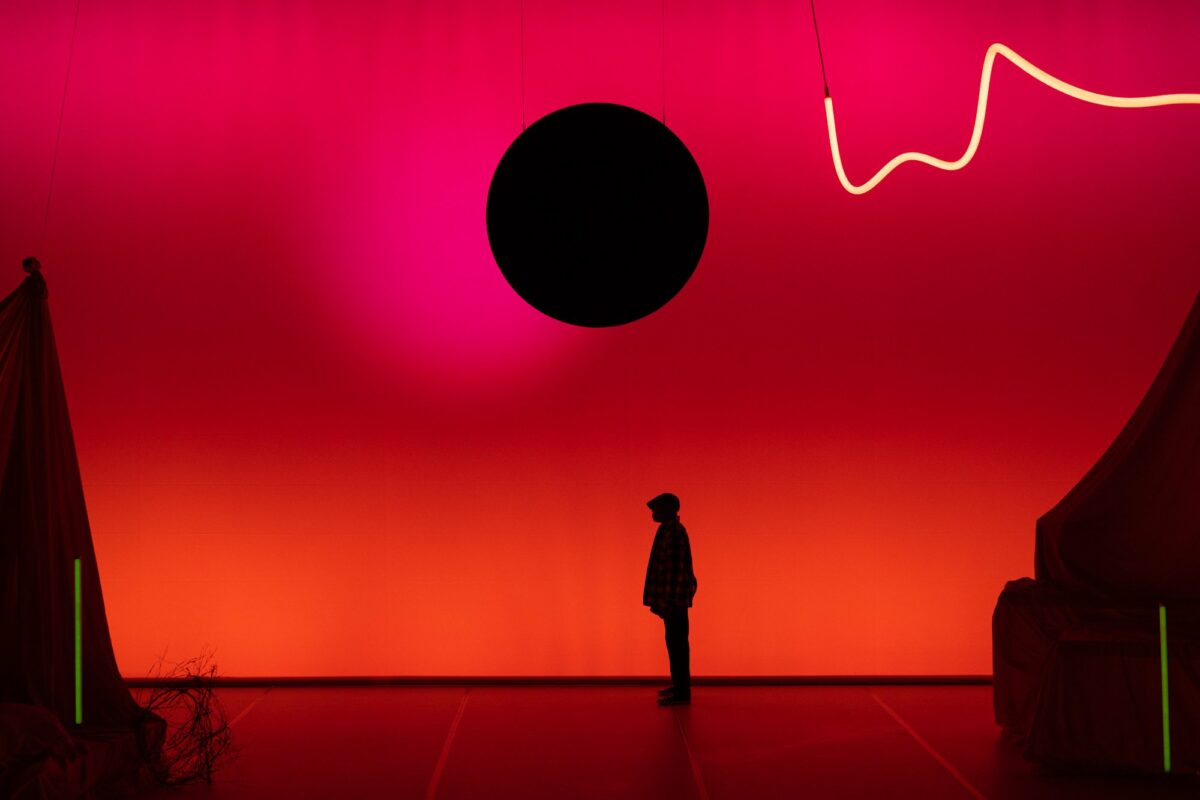 A person stands on a stage. They stand sideways on to the camera, facing left, and they are silhouetted by a glowing red backdrop. There is a silhouette of a large black circle hanging above them, and to the right  of the circle a trailing squiggle of light.