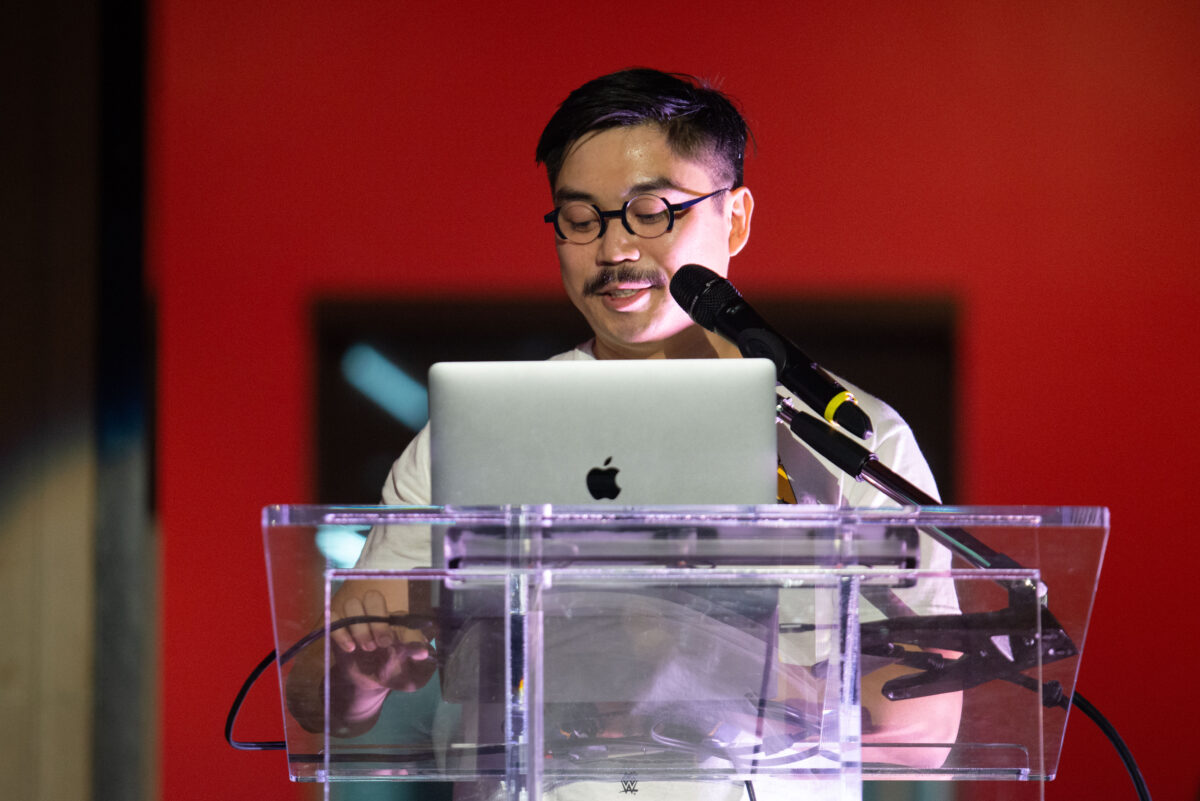 A person with dark hair, glasses and a moustache stands behind a clear acrylic podium. There is a laptop on top of the podium and a microphone partially obscuring his face. The background is dark red.