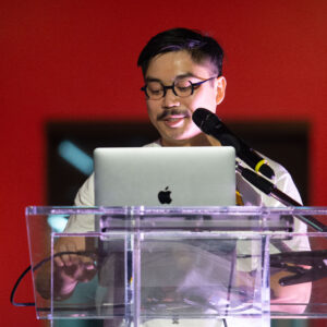 A person with dark hair, glasses and a moustache stands behind a clear acrylic podium. There is a laptop on top of the podium and a microphone partially obscuring his face. The background is dark red.
