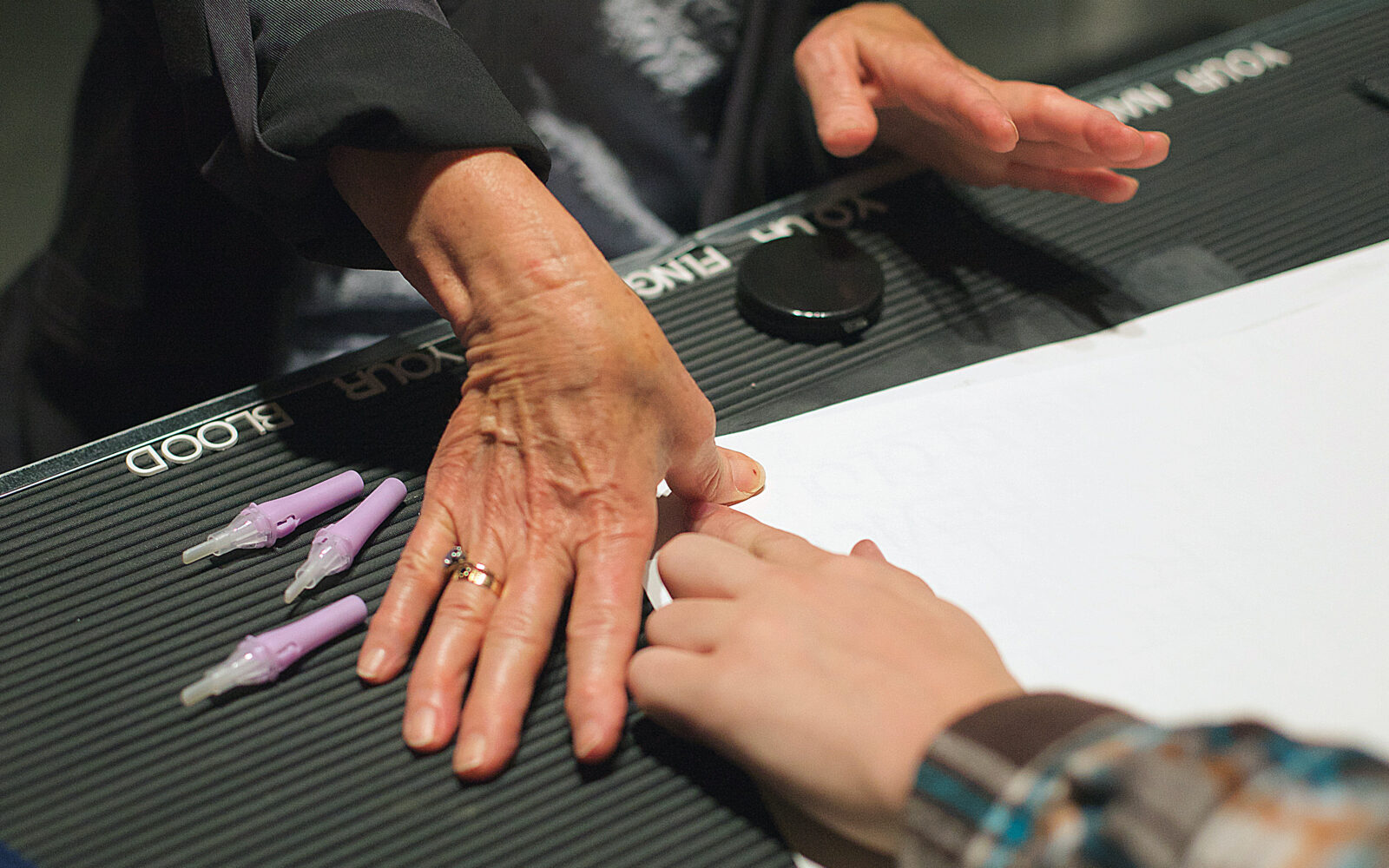 Two people touch hands across a table. One is older, one is younger. To the side of their touching hands are 3 finger prickers.