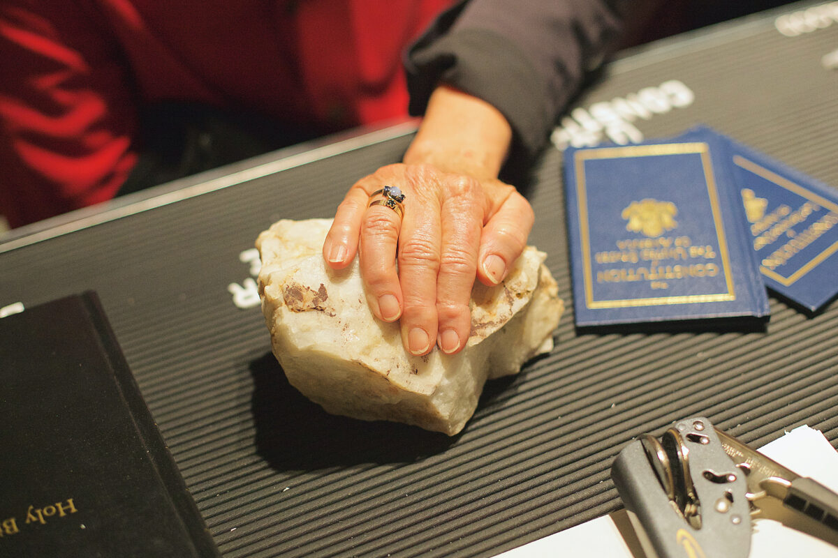 A person lays a hand on top of a rock which is on a tabletop. Next to the rock is a holy bible and the United States Constitution.