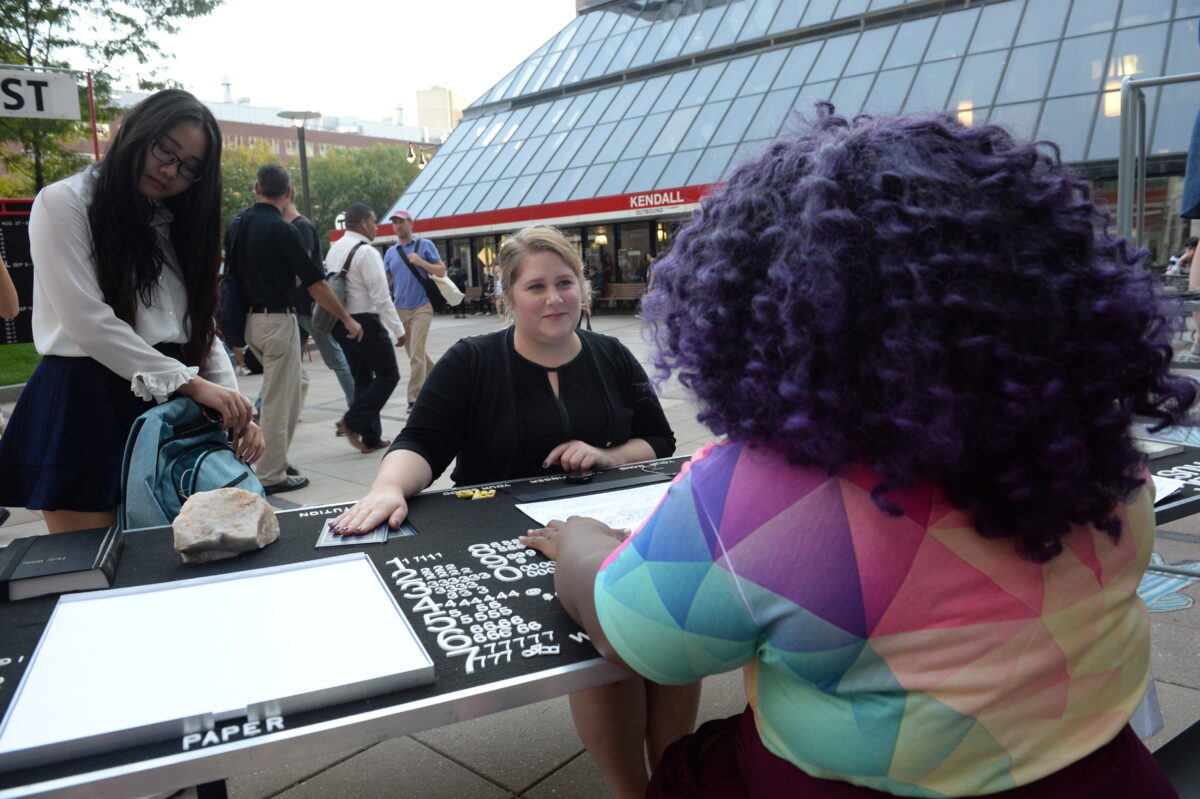 Two people sit opposite each other at a table outside a large building. The person facing the camera has their right hand on a book. There are blank pieces of paper on the table.