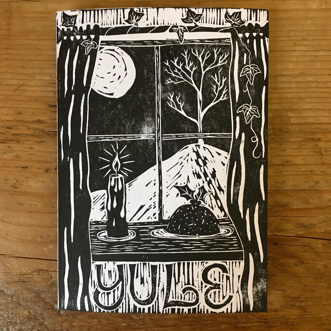 A black and white greeting card featuring a view out of a window with a Christmas putting and a candle on the windowsill. Underneath the window it says 'yule'.