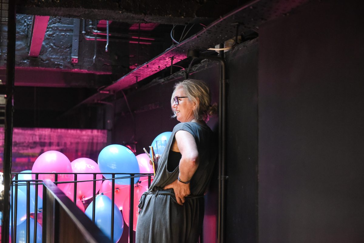 A woman leaning against a wall with pink and blue balloons in the background.
