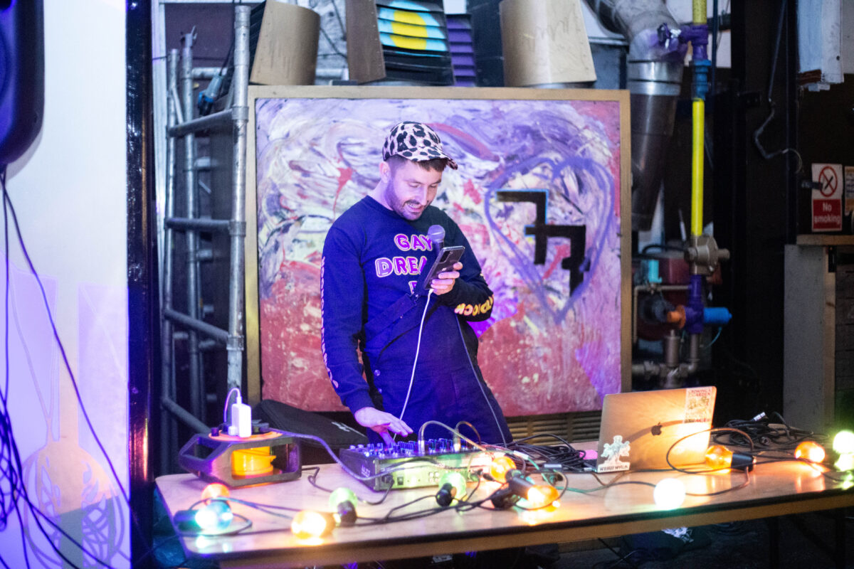 A man wearing a leopard print cap and a dark long sleeved t shirt stands behind a table. He holds a microphone and a phone in his left hand, and presses buttons on an audio mixer with his right hand. Also on the table are festoon lights, a macbook and an electric extension cable. There is an abstract art painting in the background.