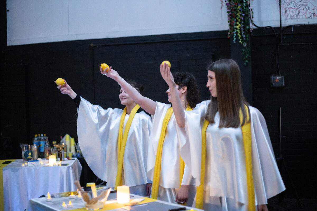 Three people wearing choir robes with yellow scarves stand in a line, each raising a lemon up in the air with their right hand. There are tealight candles on the table in front of them. Behind them is a black wall.
