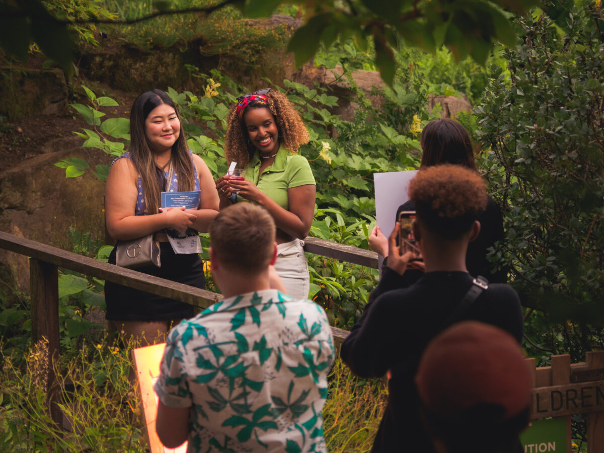 Two female-presenting people stand on a footbridge in a botanical garden. They are smiling and looking toward a person holding up a large piece of cardboard. A person in the foreground holds a phone up to take a photograph of them. Two additional people stand in the foreground, backs facing the camera.
