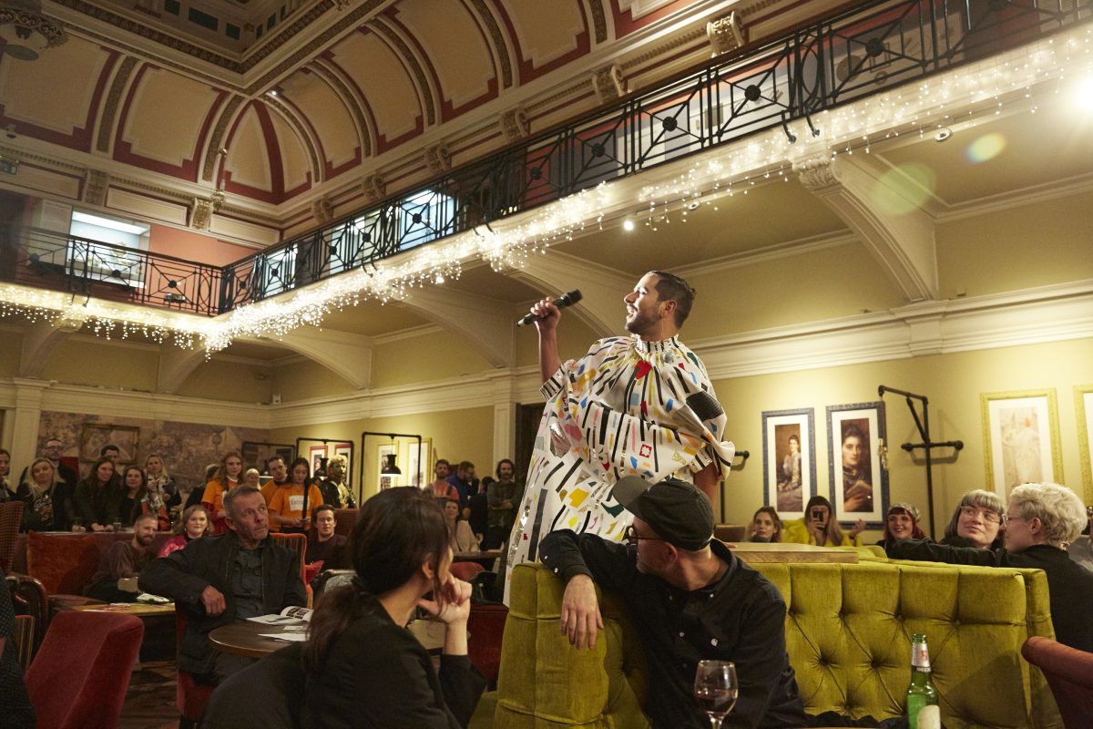 A person in a colourful outfit sings in the centre of a large, seated crowd.