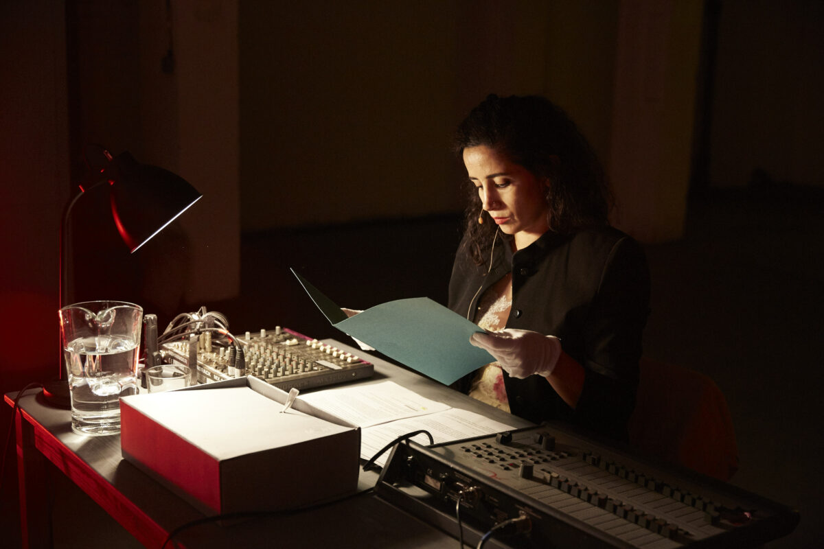 A woman sits at a desk in dim lighting. She is wearing a pair of white gloves and holding a document in her hands. On the table in front of her is a lamp, a jug of water, a box of paperwork, and two sound mixing desk controllers/