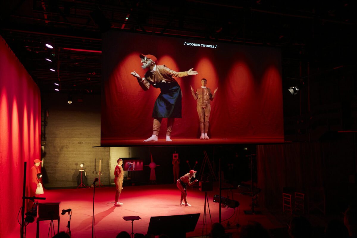 Two performed stand on a red stage, their image is projected onto a screen above them.