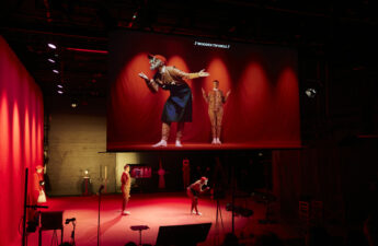 Two performed stand on a red stage, their image is projected onto a screen above them.