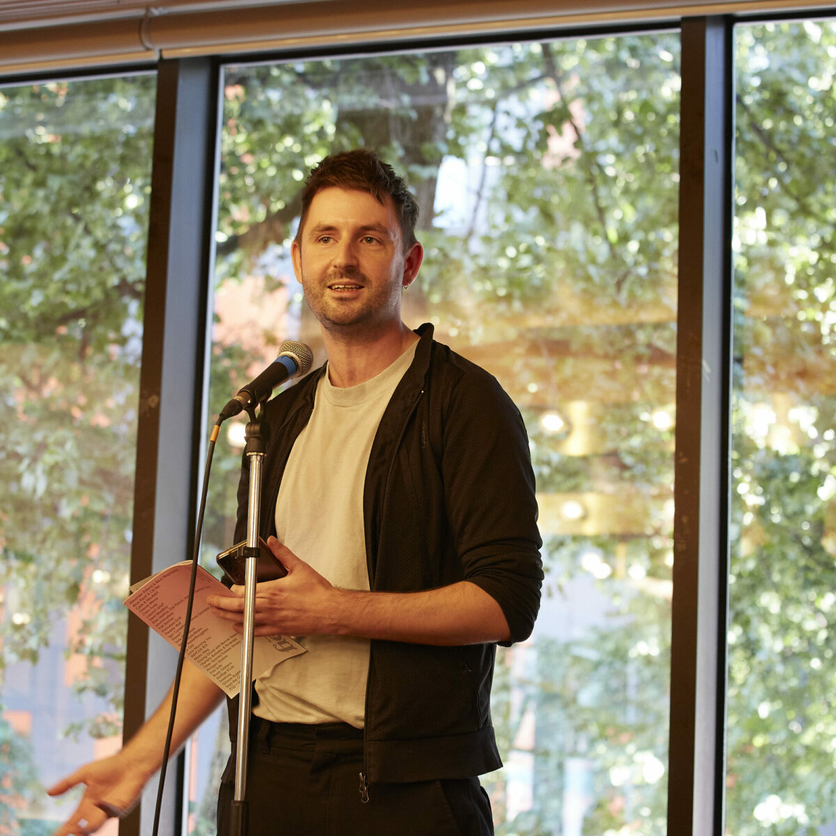 Person talking into a microphone, standing in front of a large window