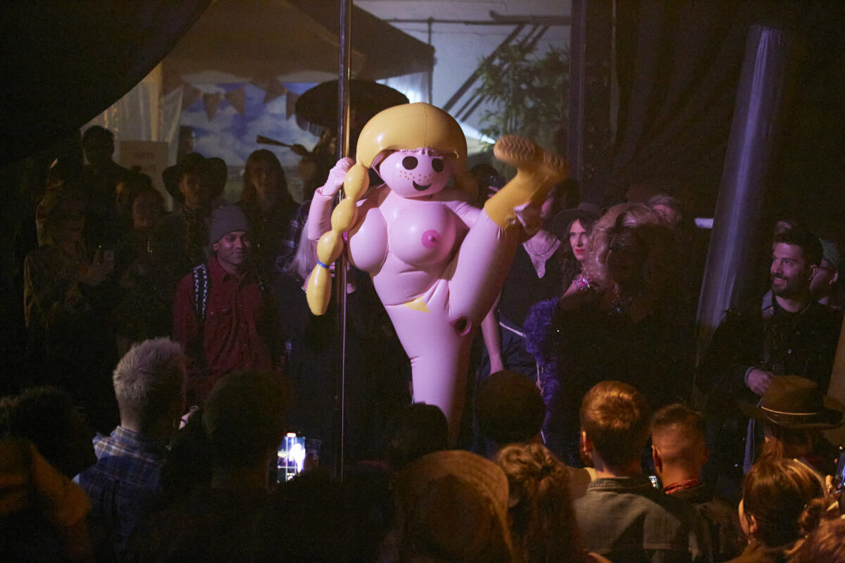 An inflatable doll performs a strip tease on a pole, inside a nightclub. The doll is surrounded by people dancing.