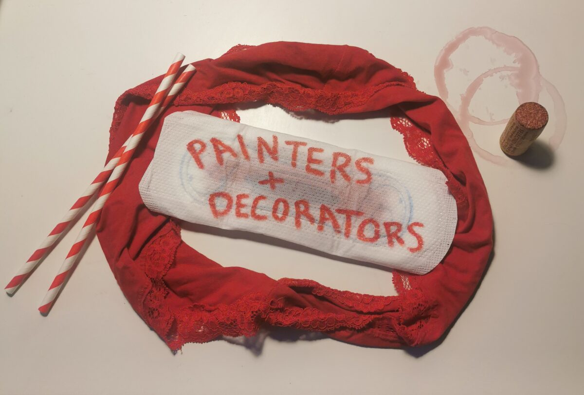 A soiled panty liner inside a pair of red knickers. There are red words on the liner that read: painters + decorators. Two red and white striped straws lie to the left of the knickers. two red wine rings stain the surface to the right of the knickers. There is a cork from a wine bottle next to them. 
