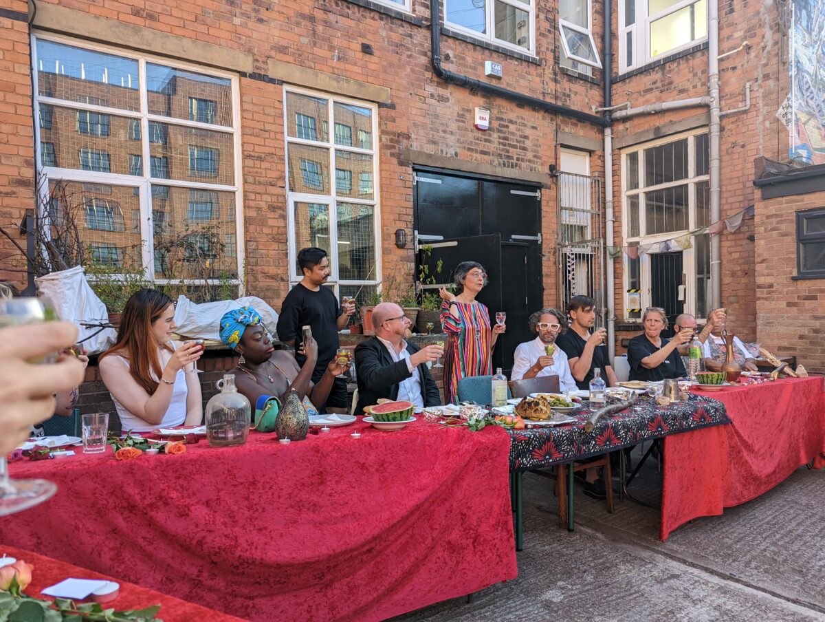 10 people sat at a long banquet table raise a glass to make a toast. The table is covered by a red table cloth, and there are plates of food and jugs of water across the length of it. The table is outside, with a red brick building in the background.