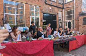 10 people sat at a long banquet table raise a glass to make a toast. The table is covered by a red table cloth, and there are plates of food and jugs of water across the length of it. The table is outside, with a red brick building in the background.