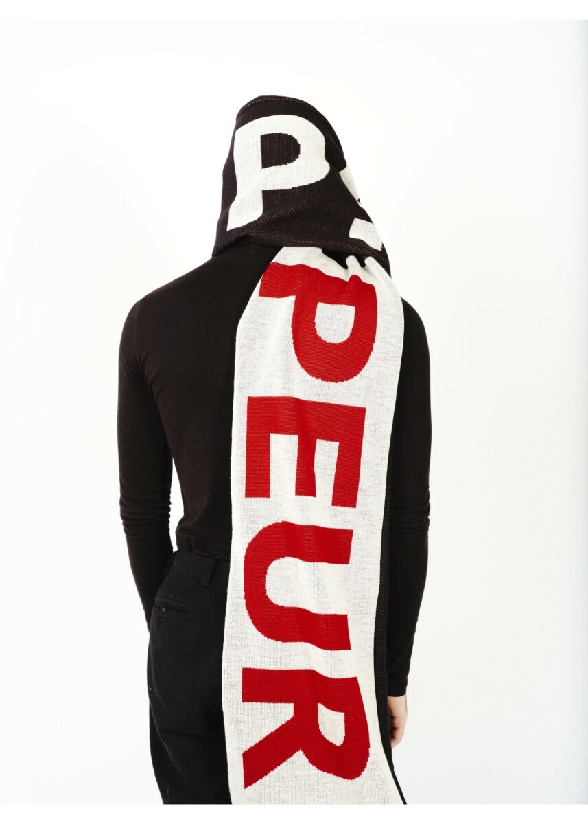 A person dressed in black has a large scarf wrapped around their head and hanging down their back. The scarf has the letters PPEUR visible. 