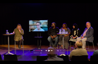 5 people on stage sitting on stools. A large screen shows a headshot of someone joining the talk remotely over video call.