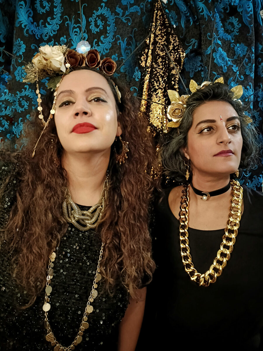 Two women stand in front of a dark floral background. They are both adorned with gold jewellery and floral head dresses. 