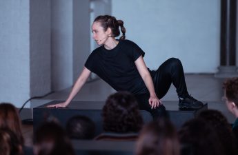 Lisa sits on a stage dressed in all black with one leg slightly raised and bent at the knee and one leg beneath her on the floor.