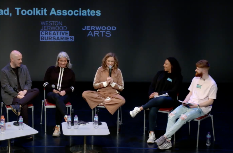 Five people sit side by side on a stage as part of a panel discussion. There are two low coffee tables in front of them with water bottles on. The backdrop is black with the words 'Weston Jerwood Creative Bursaries' and 'Jerwood Arts' in blue.