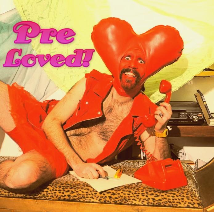 A man lies on his side, holding a red rotary phone in his left hand and a pen in his right hand. He wears red denim cut off shorts and a red biker waistcoat. His face is painted red and he has a heart shaped head piece surrounding his face. Text overlaid reads: Pre Loved!