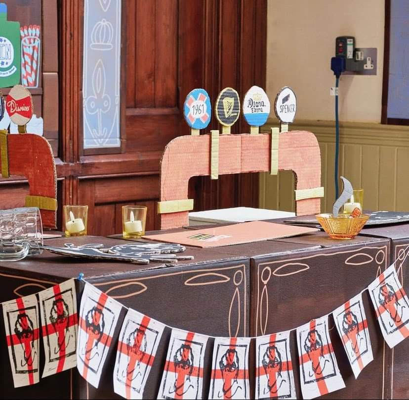 A set of beer pumps made out of cardboard, behind a bar area. Bunting of England flags featuring Princess Diana's face are draped across the front of the bar.